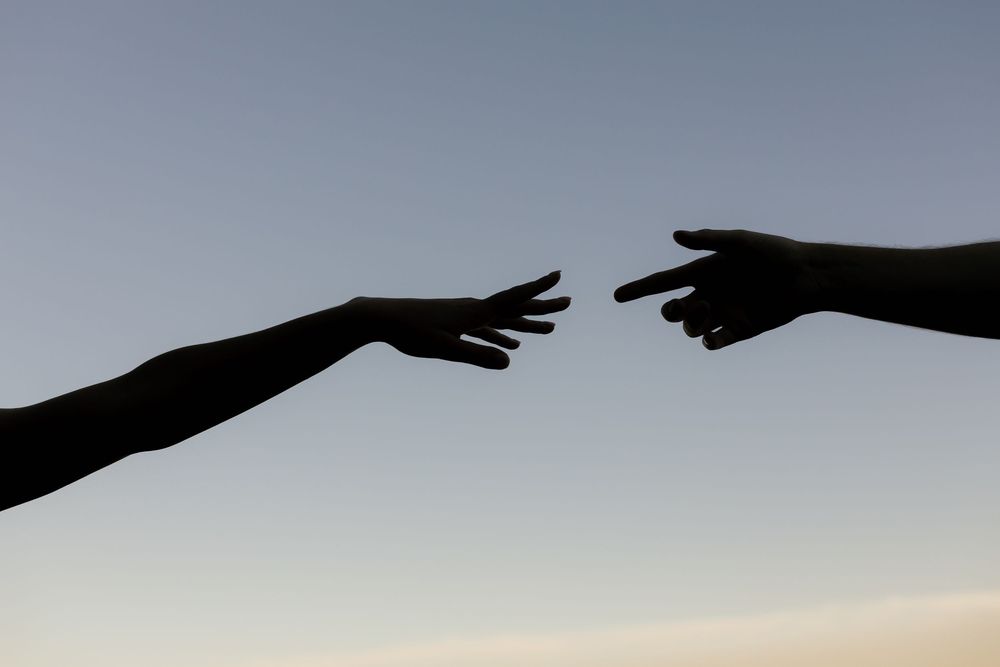 Silhouetted hands reaching across a bright sky with wispy clouds create a dramatic visual composition.