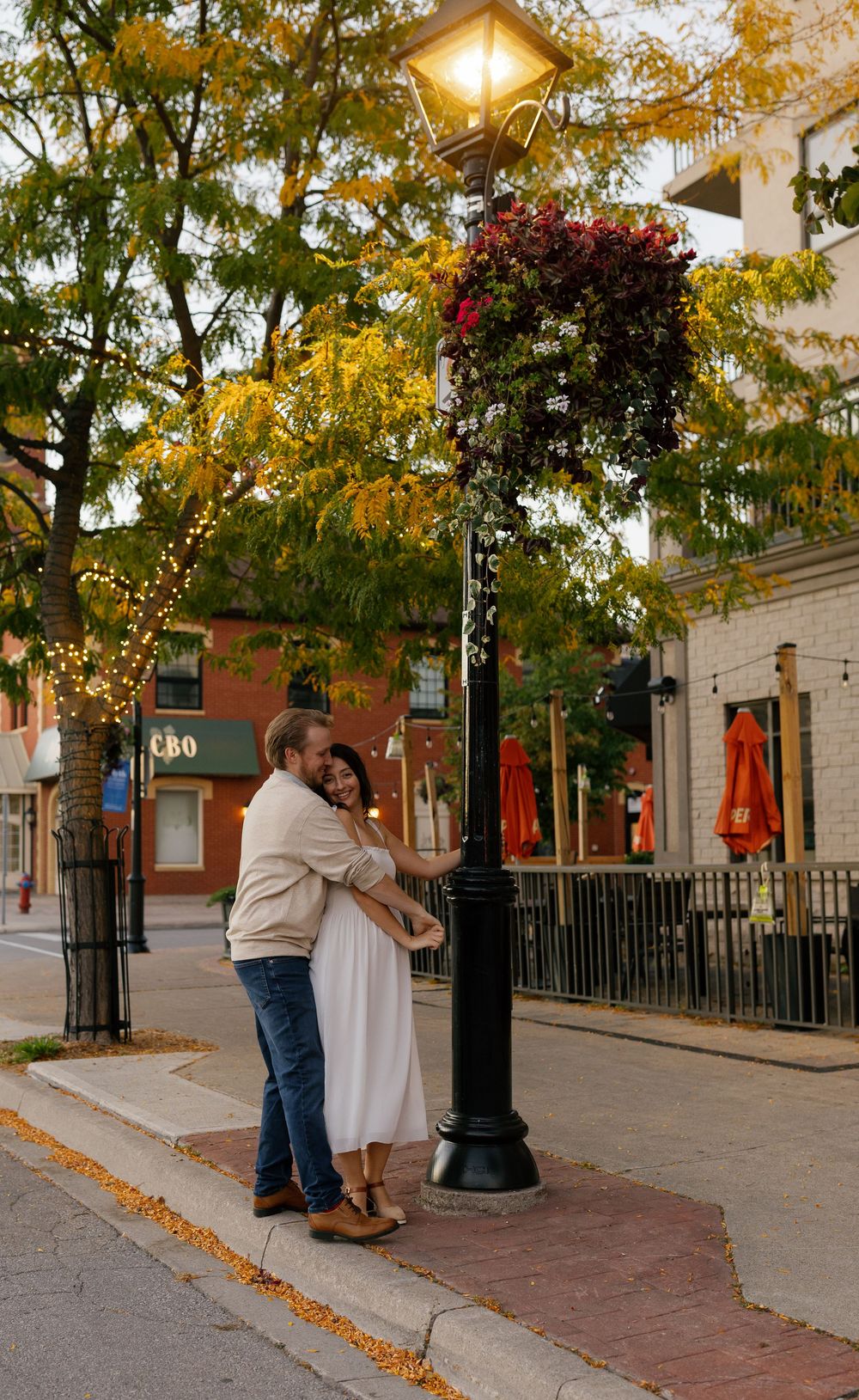 A romantic couple embraces under glowing street lamps on a brick sidewalk with autumn trees and historic buildings.