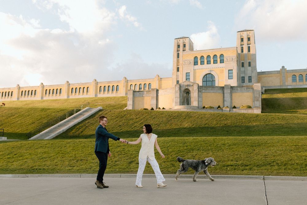 Two people walking their dogs on a grassy hill near a historic stone building on a cloudy day.