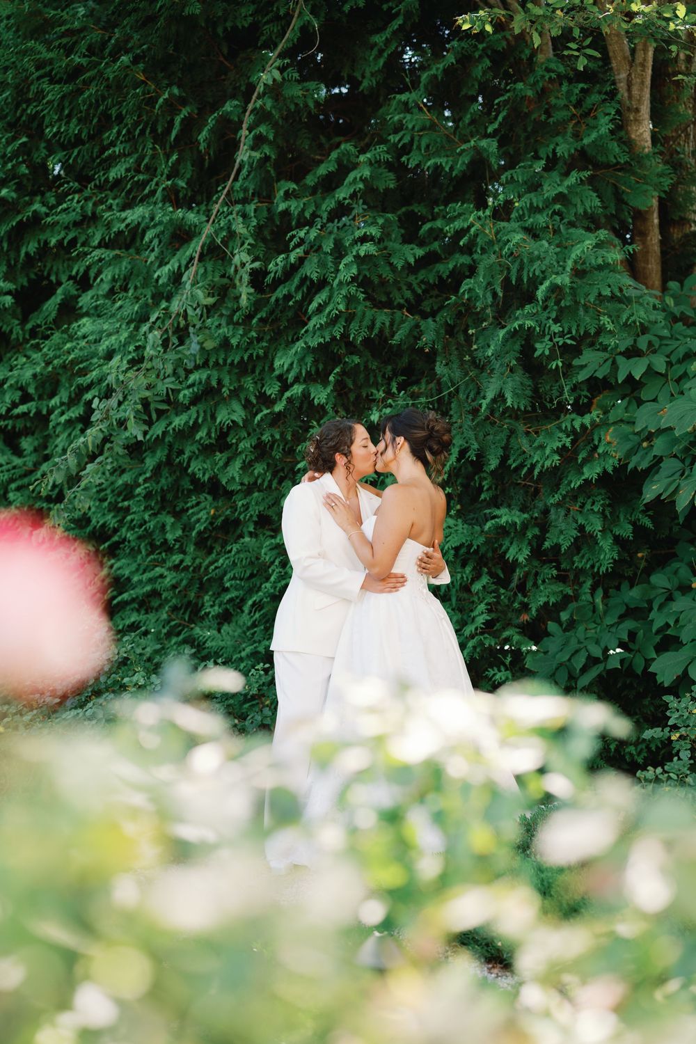A romantic couple in white outfits shares a tender kiss in a lush garden setting with green foliage in the background.