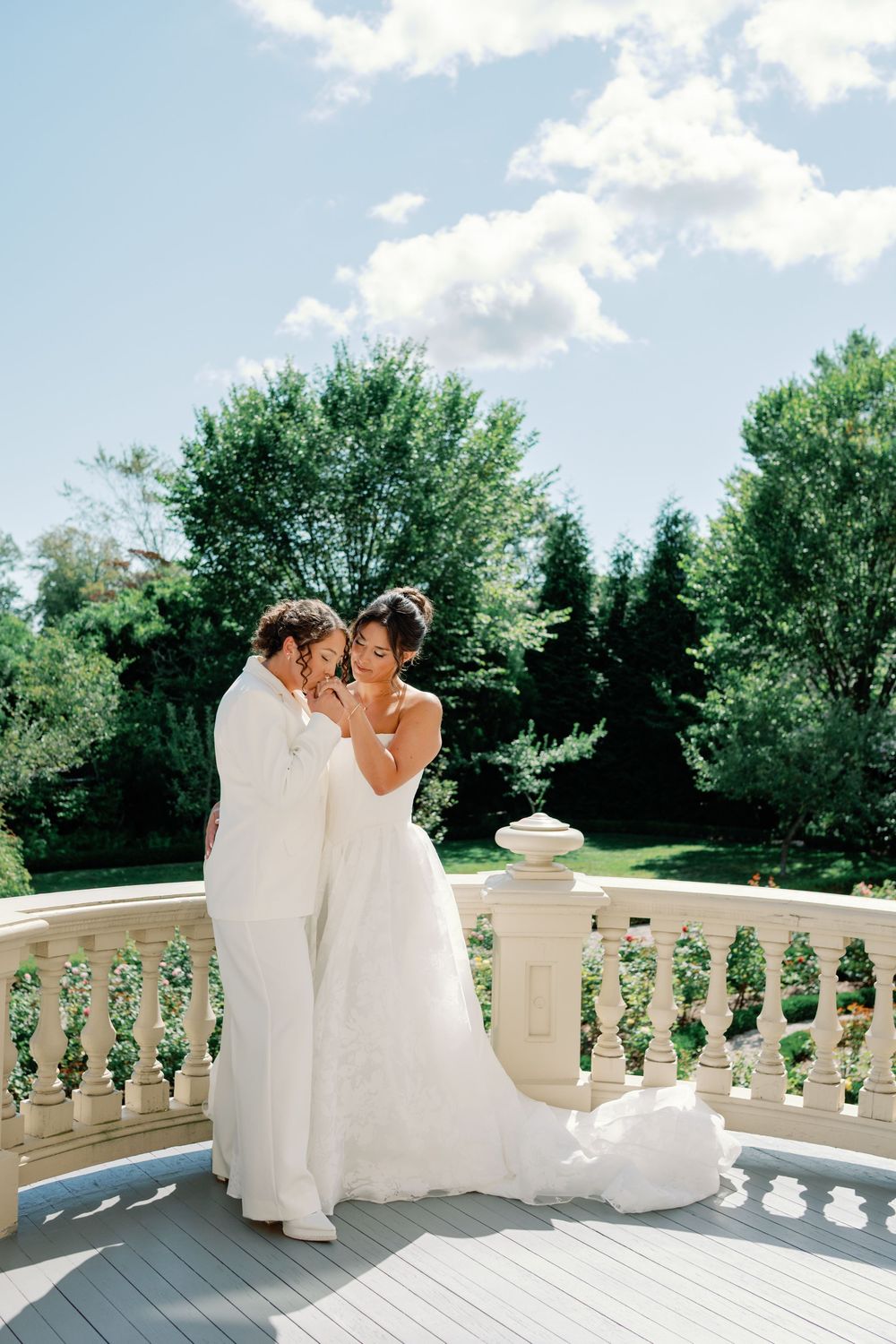 A couple in white wedding attire share an intimate moment on an elegant stone balcony surrounded by lush greenery.