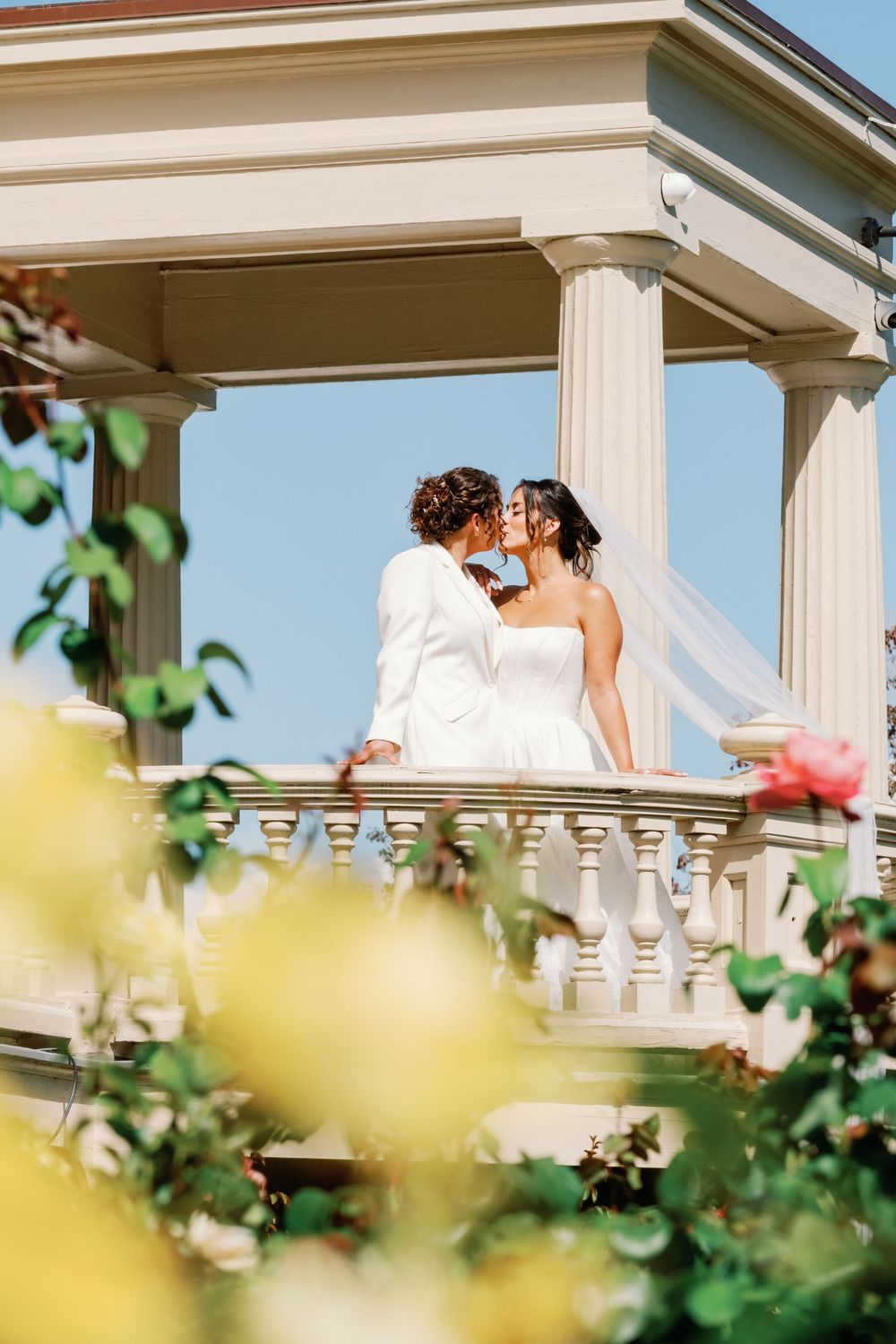 A romantic wedding couple shares tender moments on an elegant white columned balcony framed by soft greenery.