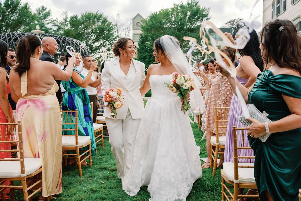 Wedding party helps bride with dress on outdoor lawn during summer celebration.