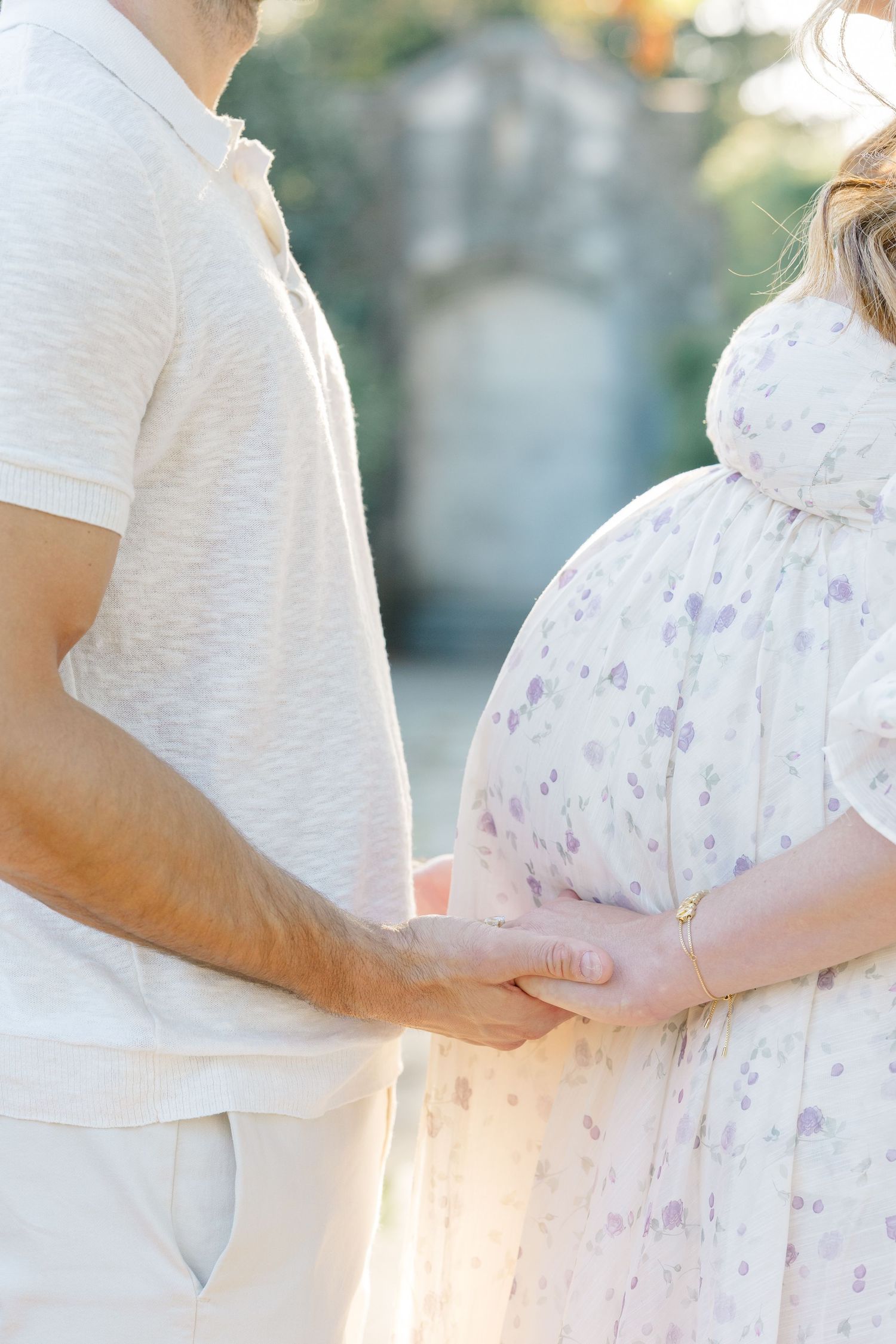 Couple in white and floral clothing embrace during maternity photoshoot at sunset in soft natural lighting.