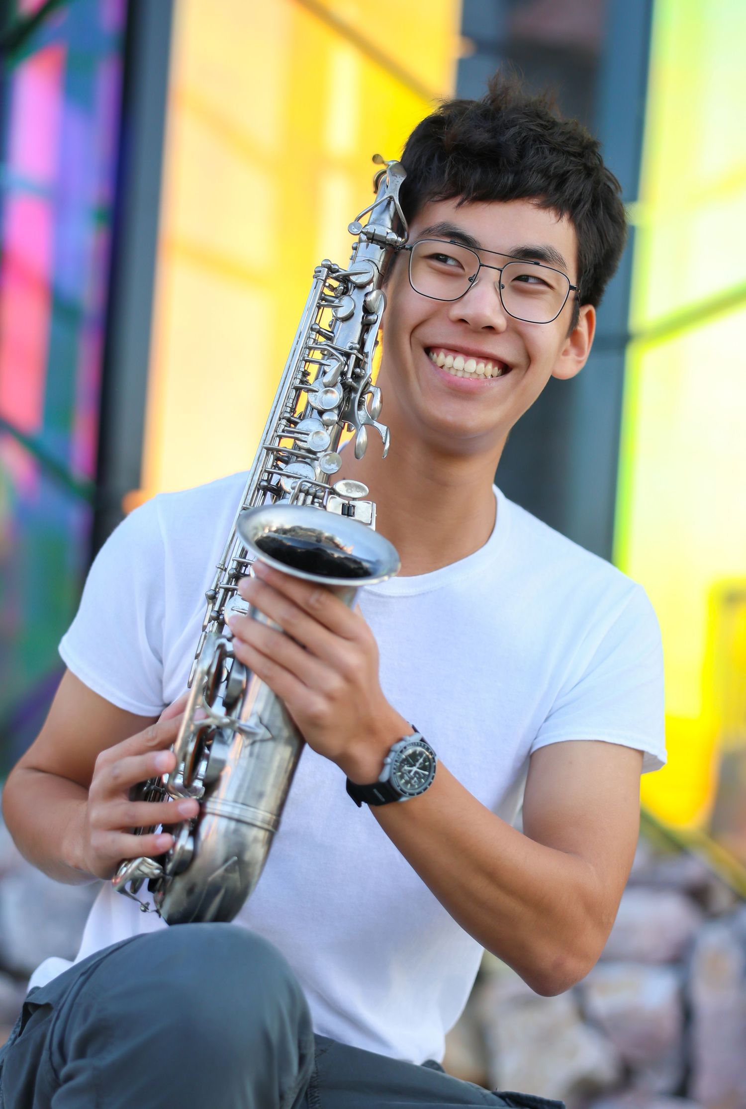 Musician poses with a silver saxophone against a colorful yellow and pink background.