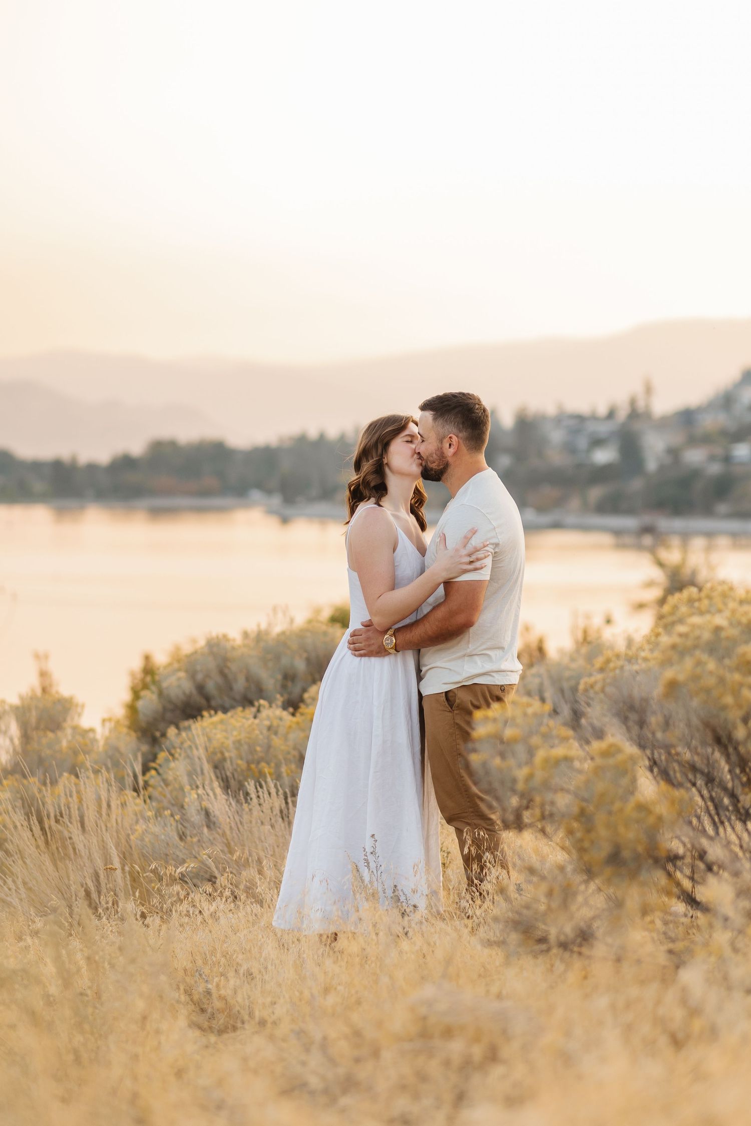 A couple embraces in a golden field at sunset with water and mountains in the background.