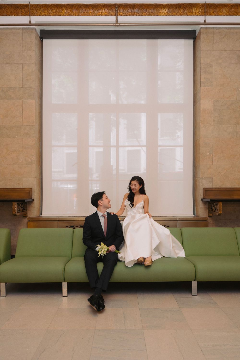 A bride and groom sit together on a modern green couch in an elegant minimalist setting.