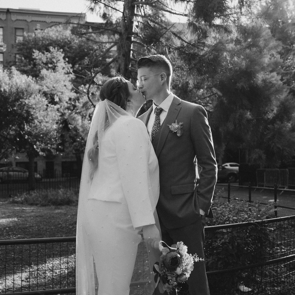 A couple poses together in a park setting with iron fencing and trees in the background.