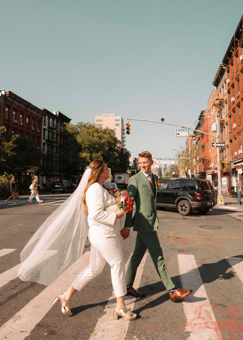 Newlyweds playfully crossing a sunny urban street with brownstone buildings in the background.