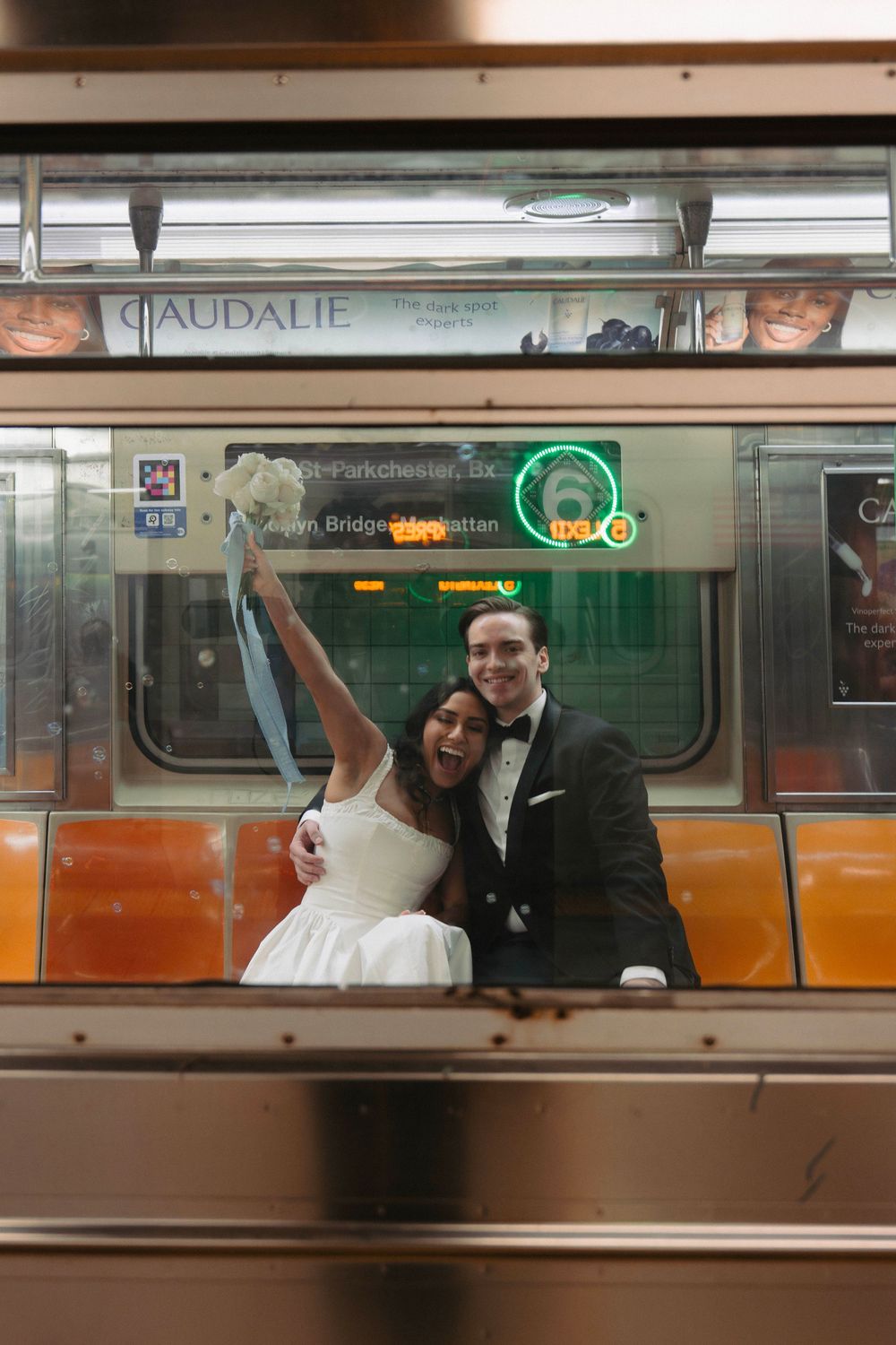 A couple in wedding attire pose joyfully inside a subway car, with the bride in white and groom in dark suit.