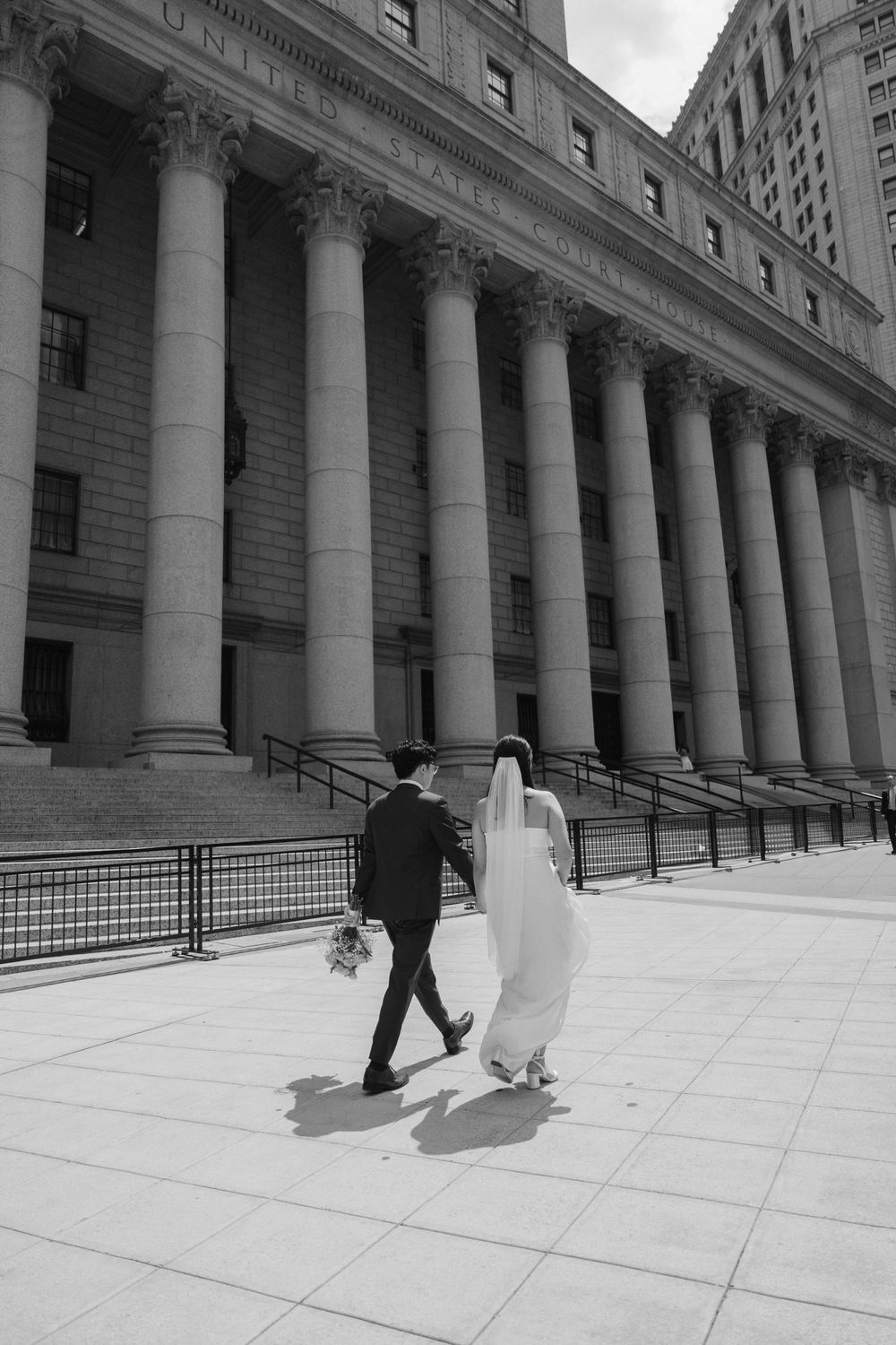 A couple in wedding attire walking up the steps of a grand courthouse building with classical columns in black and white.