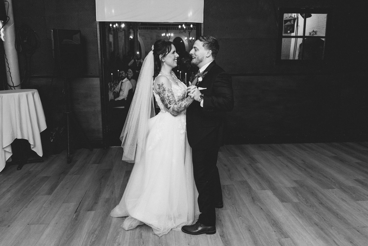 A wedding couple dances together in a dimly lit ballroom with wooden floors and romantic lighting.