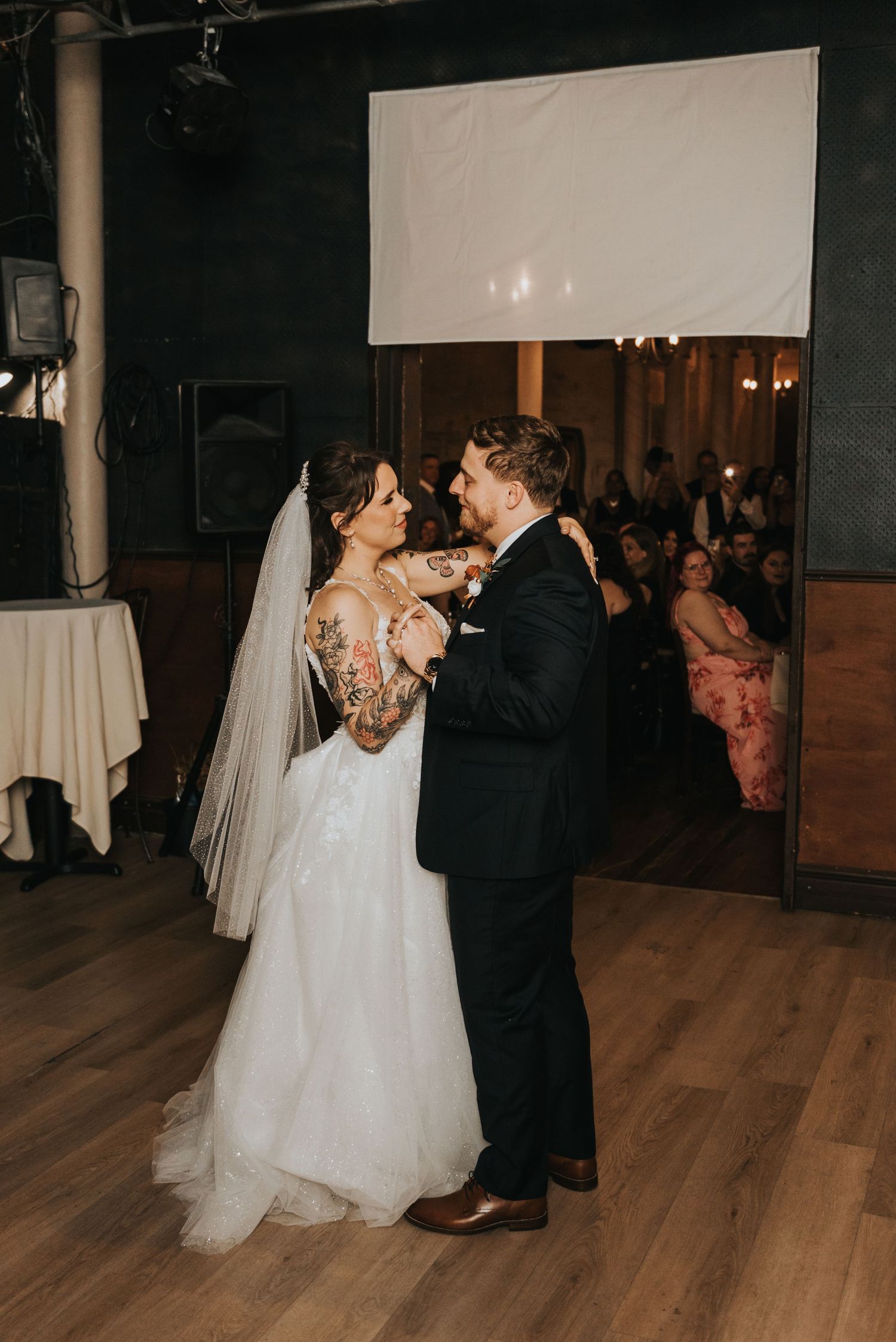 A couple shares their first dance at their wedding reception on a hardwood floor with a white projection screen in background.