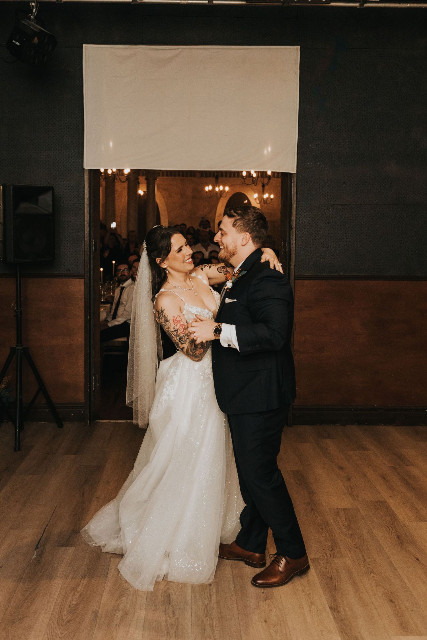 A couple embraces during their wedding dance on a wooden floor with warm lighting and a projection screen behind them.