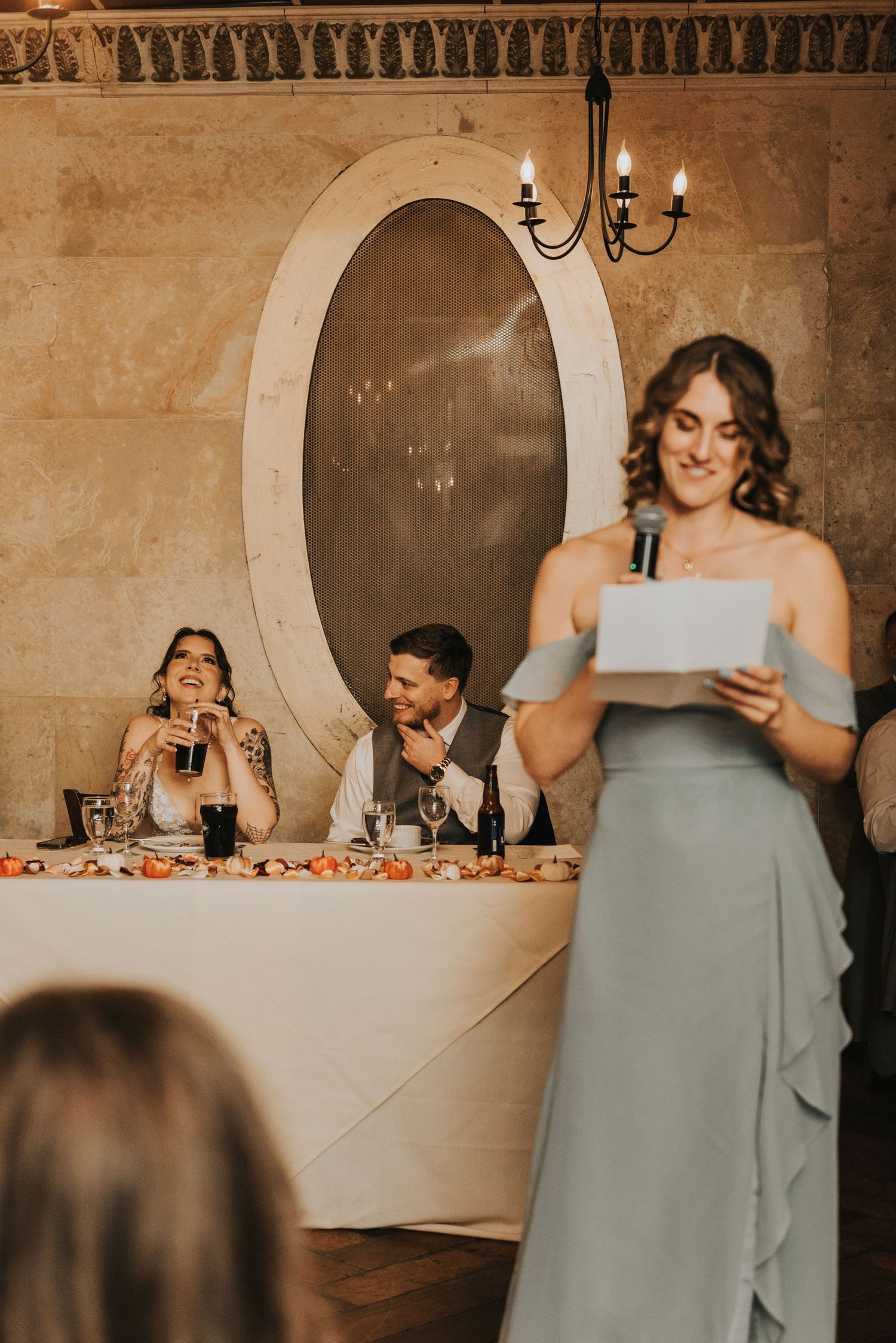 Wedding party members in light blue dresses deliver emotional speeches at an elegant stone-walled reception venue.