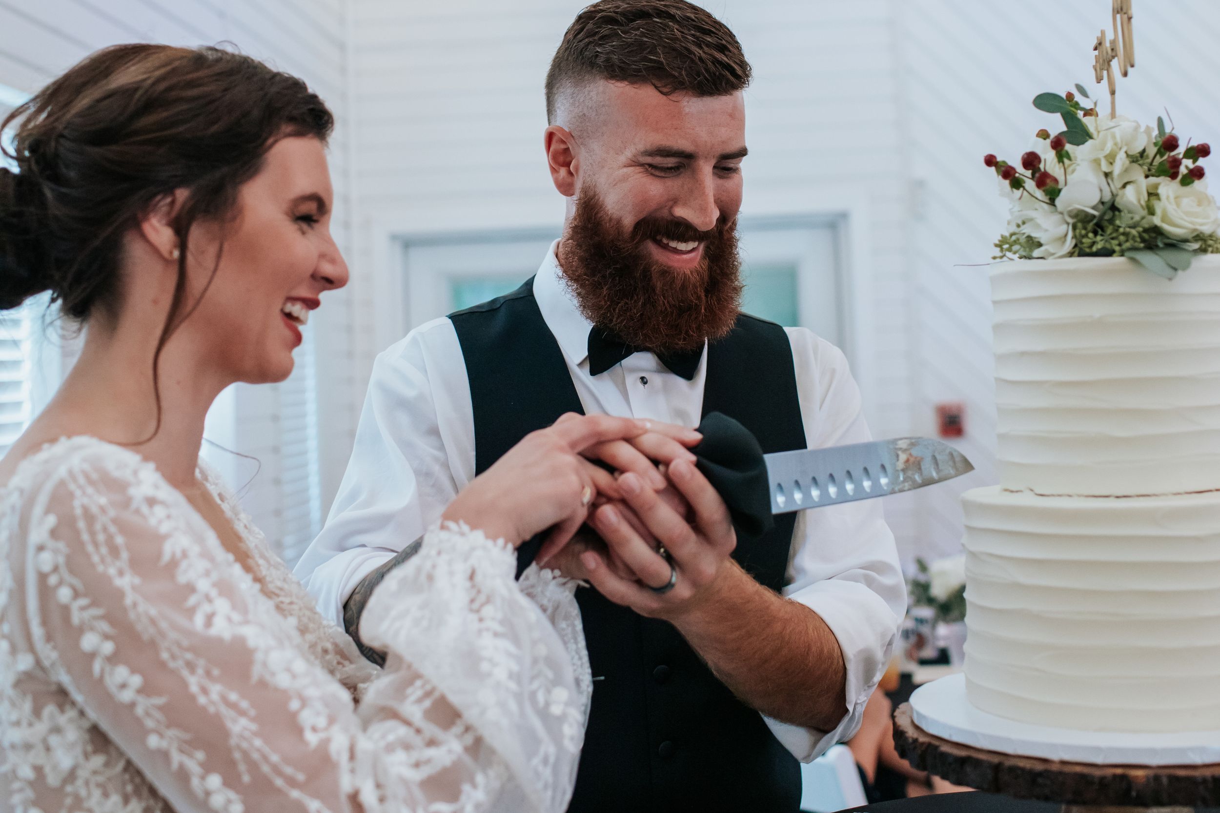 Wedding cake cutting ceremony in a bright white venue with string lights in the background.