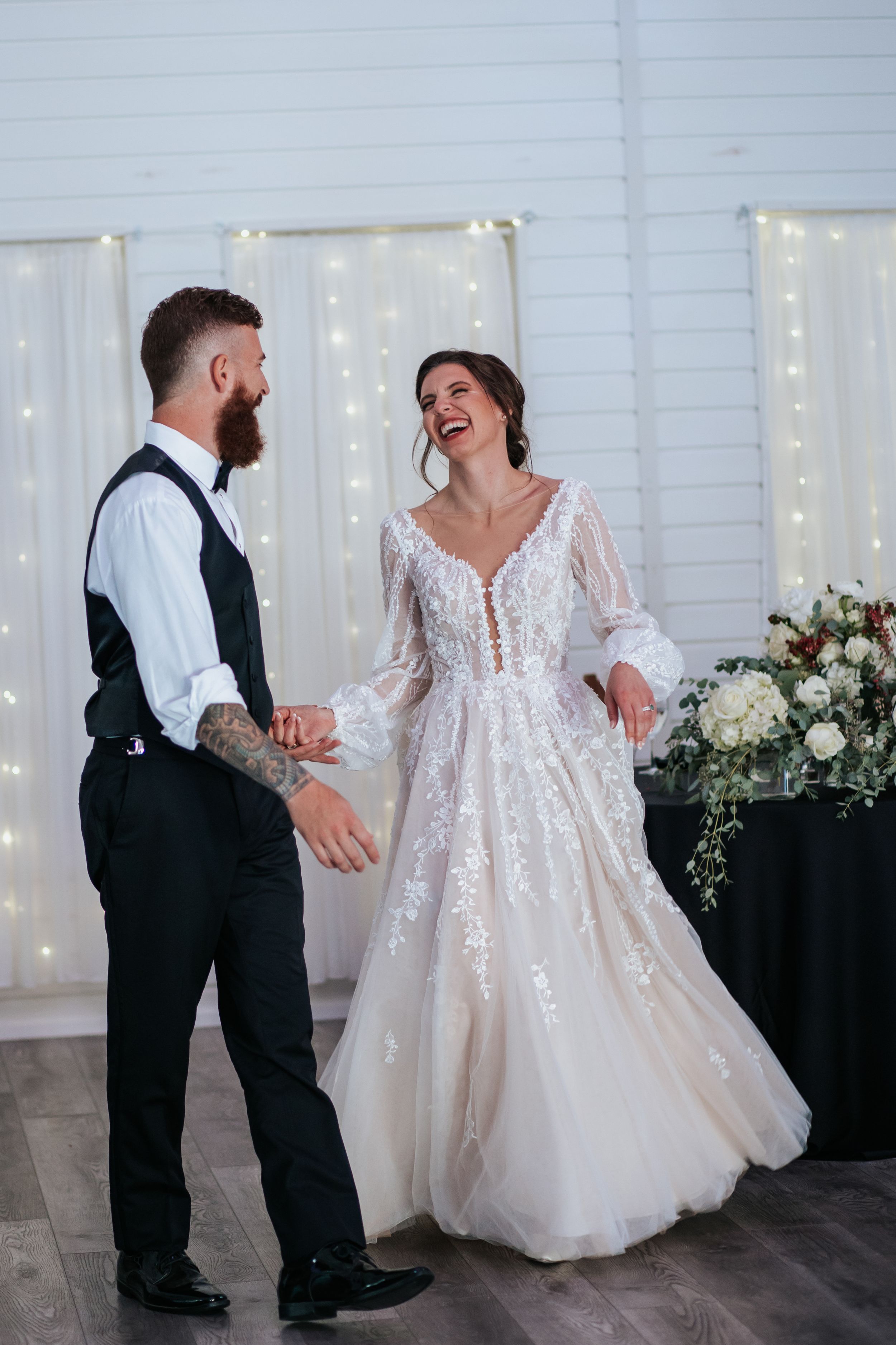 A playful wedding dance moment captured in a white-walled venue with twinkling lights.