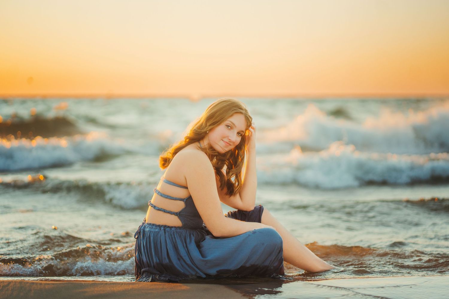 Teen girl in blue outfit sitting on rocks at sunset with ocean waves crashing behind her.