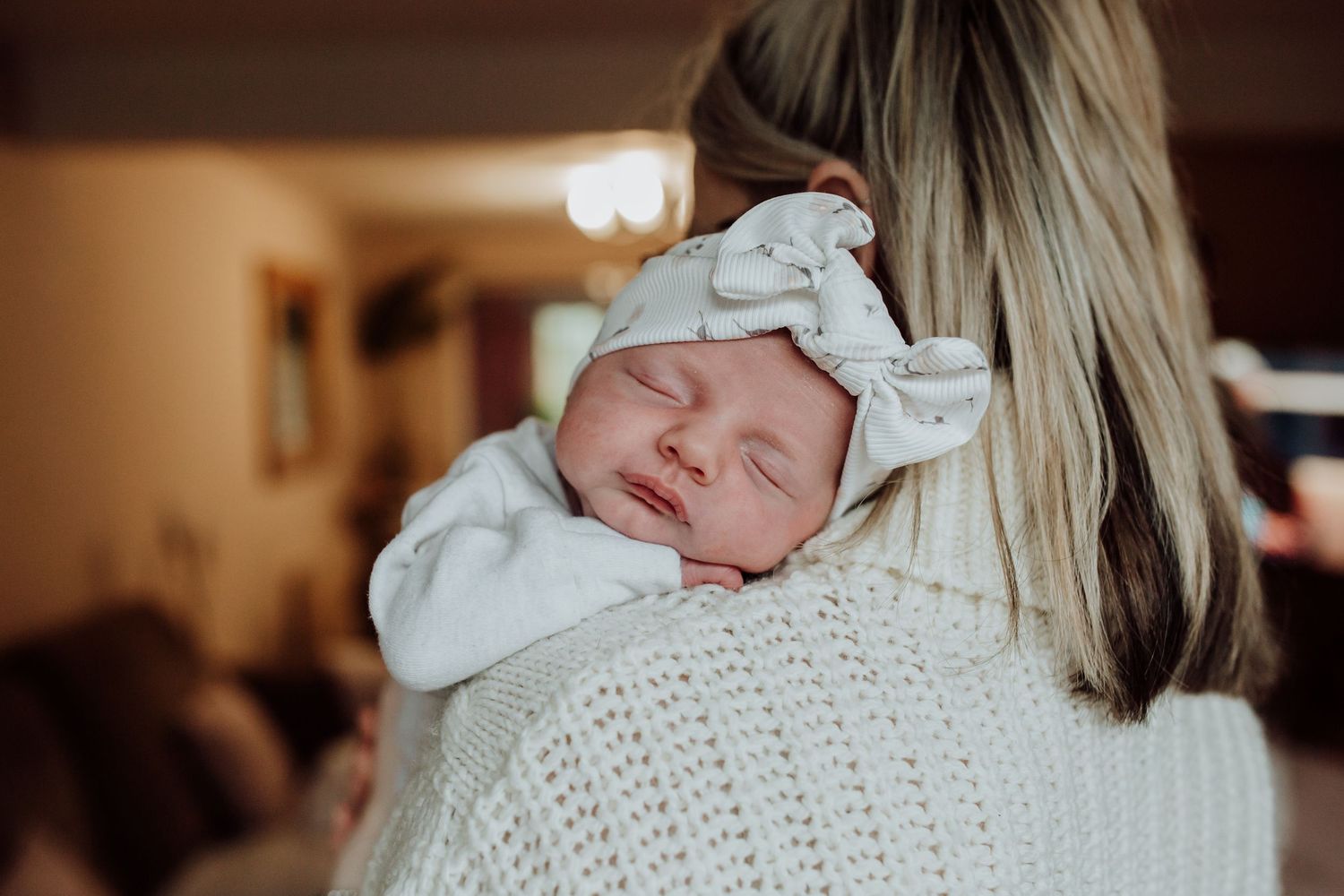 Newborn baby wearing white bow headband sleeps peacefully while being held in warm lighting. Lifestyle Newborn photography