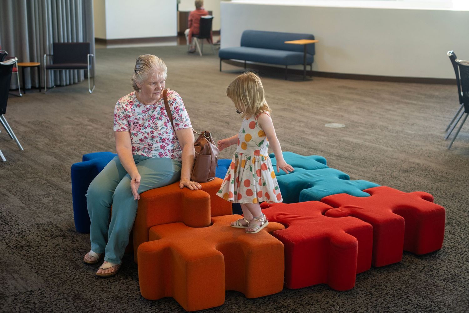 Grandmother and toddler girl playing on colorful puzzle-shaped foam seats in a modern indoor play area.