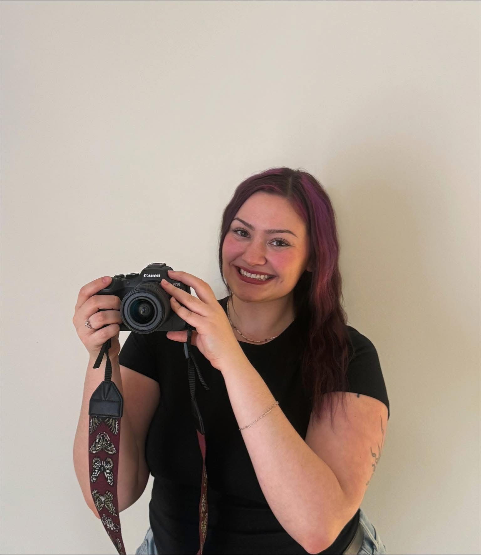 Smiling woman with purple hair holding a Canon camera with a decorative strap against a white wall.