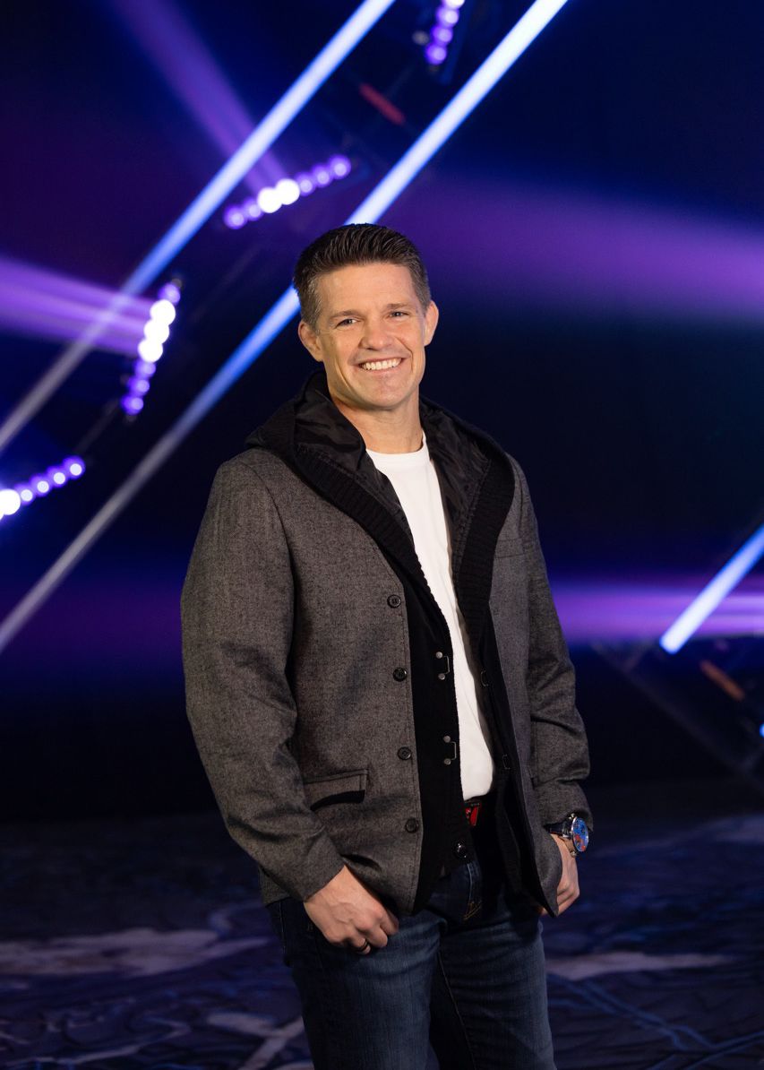 Russell Brunson smiling in gray jacket and jeans poses confidently against vibrant purple and blue stage lighting backdrop. FHL headshot
