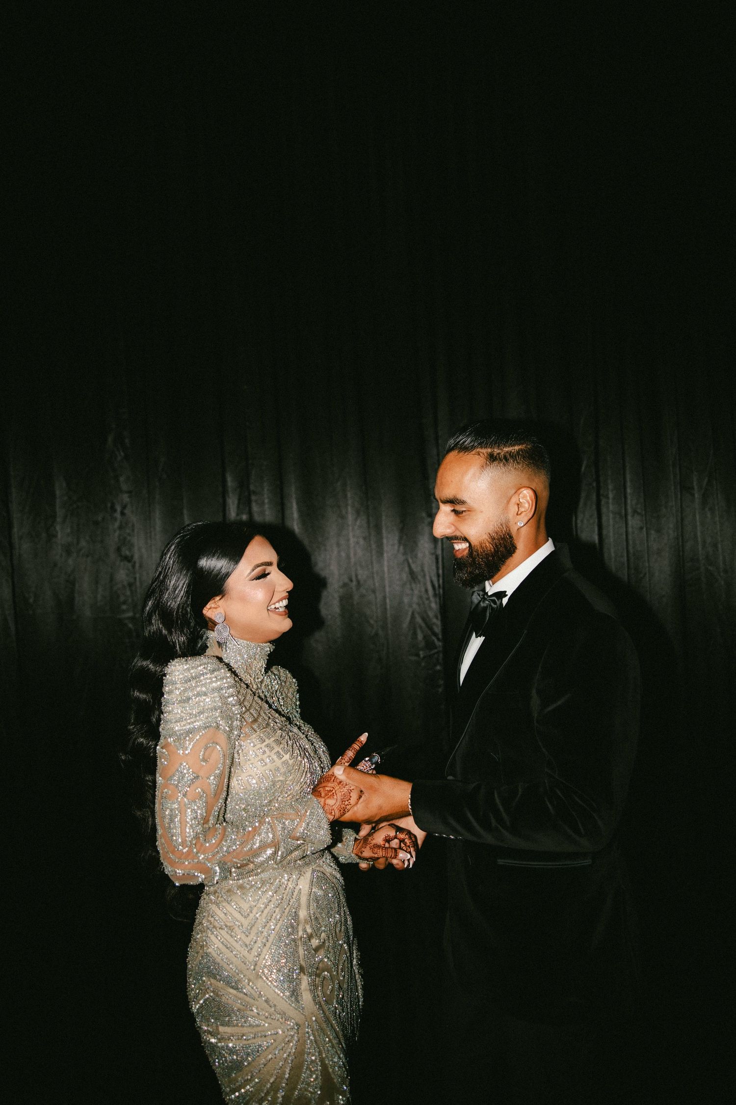 Elegant couple in formal attire holding hands, woman in beaded gown and man in black velvet tuxedo, against dark backdrop.