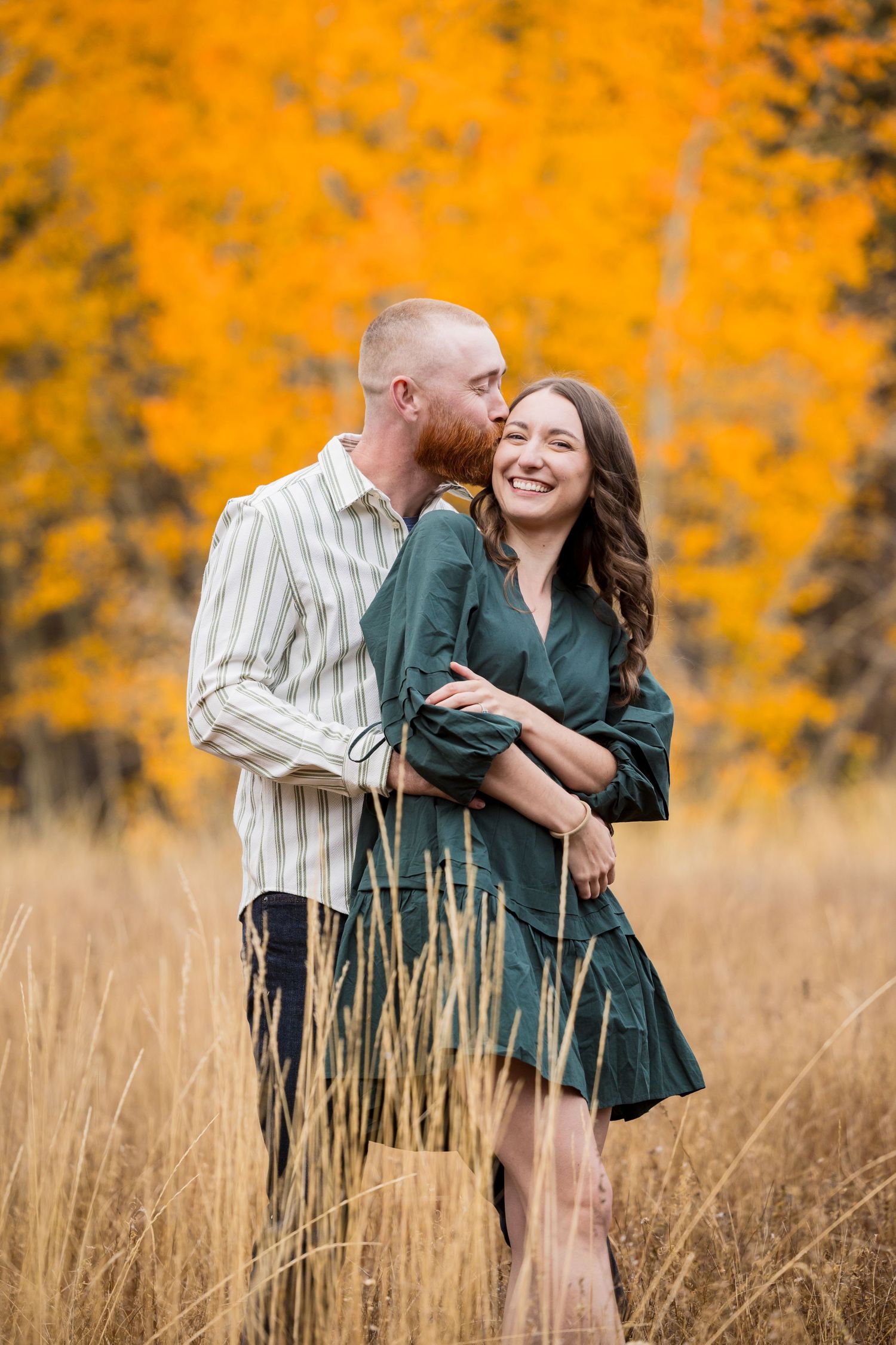 Couple embracing in autumn field with golden aspen trees, man kissing woman's cheek as she smiles joyfully.