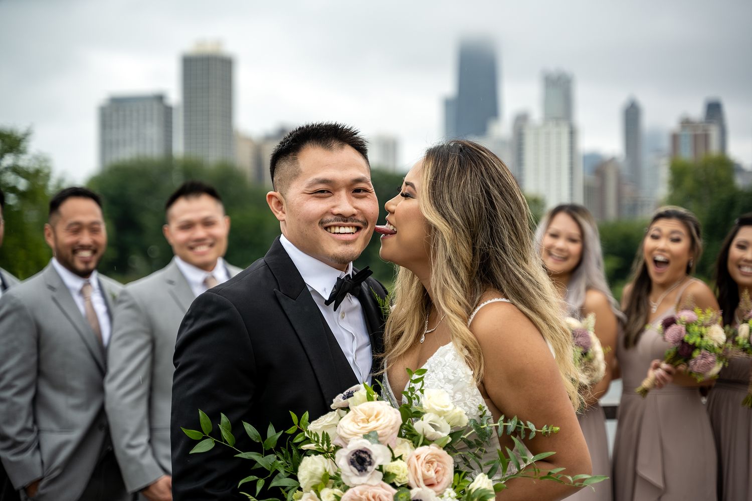 Bride kisses groom on cheek at outdoor Chicago wedding with city skyline backdrop and smiling wedding party.