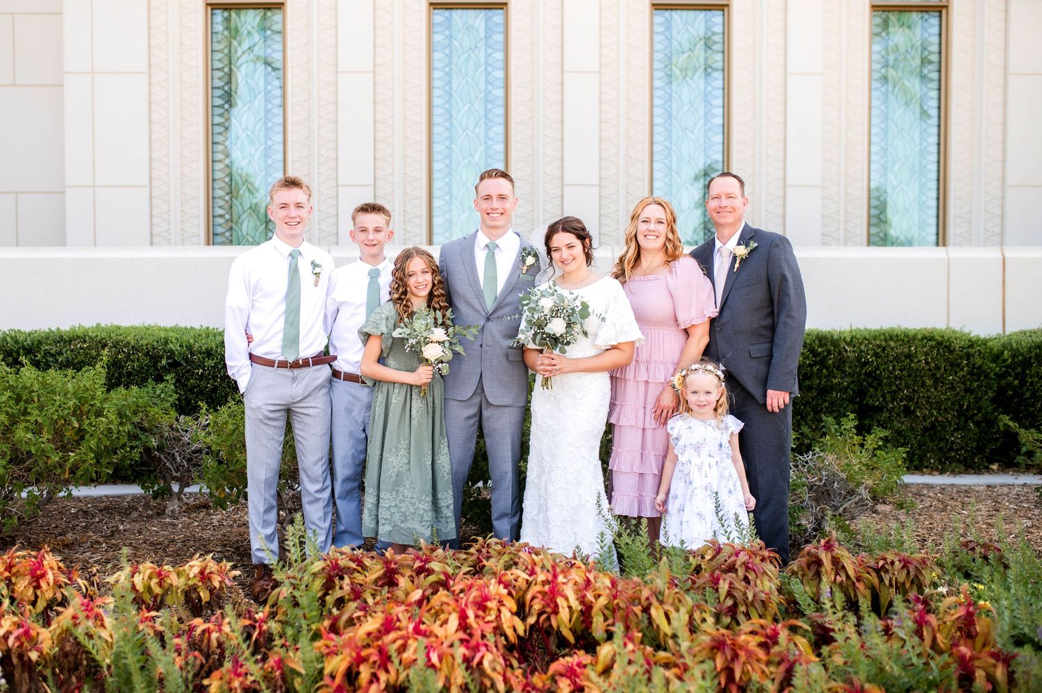 Wedding party family portrait outside an LDS temple surrounded by lush greenery and flowers.