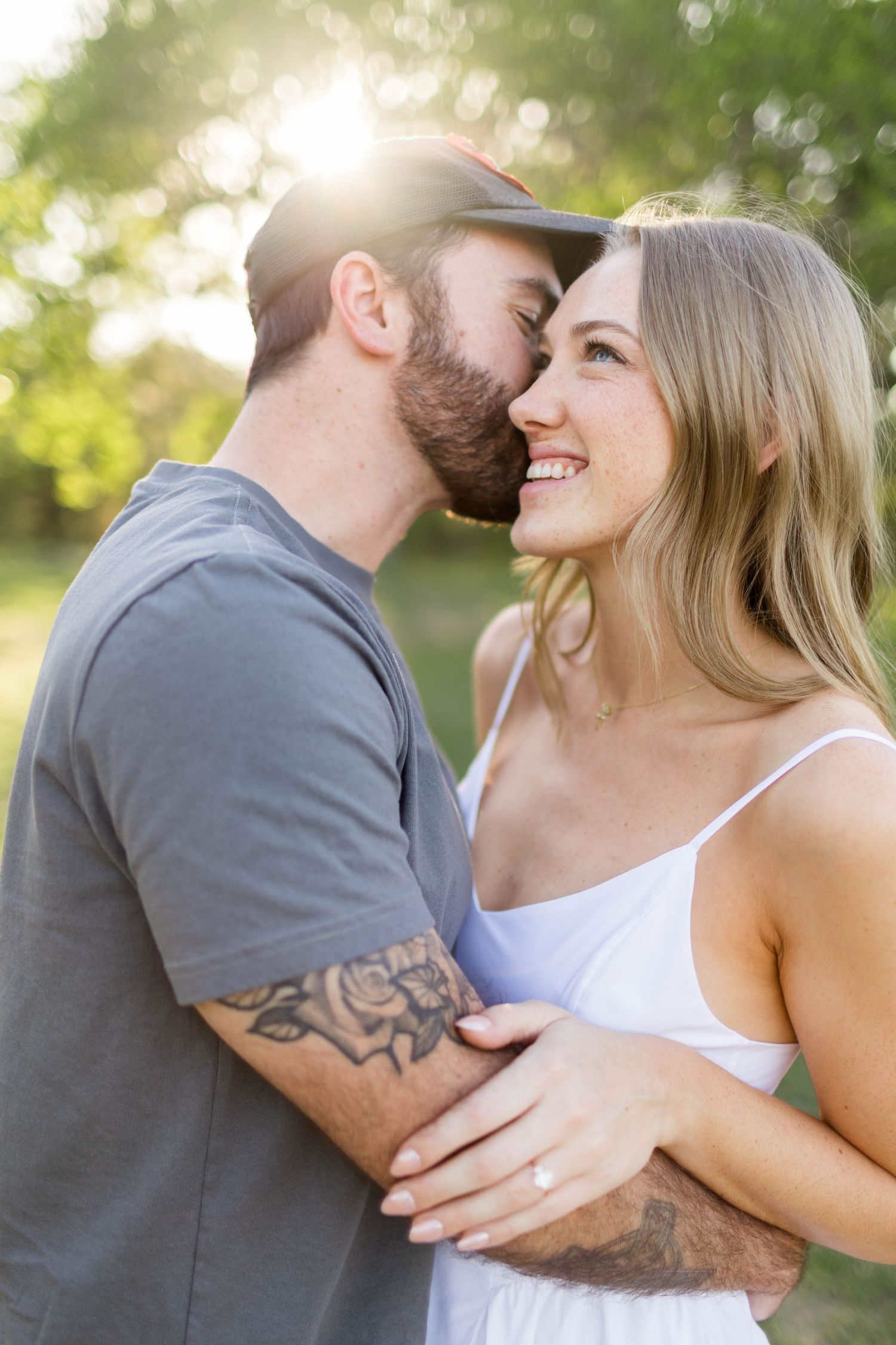 A couple shares a romantic moment outdoors, man with tattoos and cap kissing smiling blonde woman in white dress in Austin.
