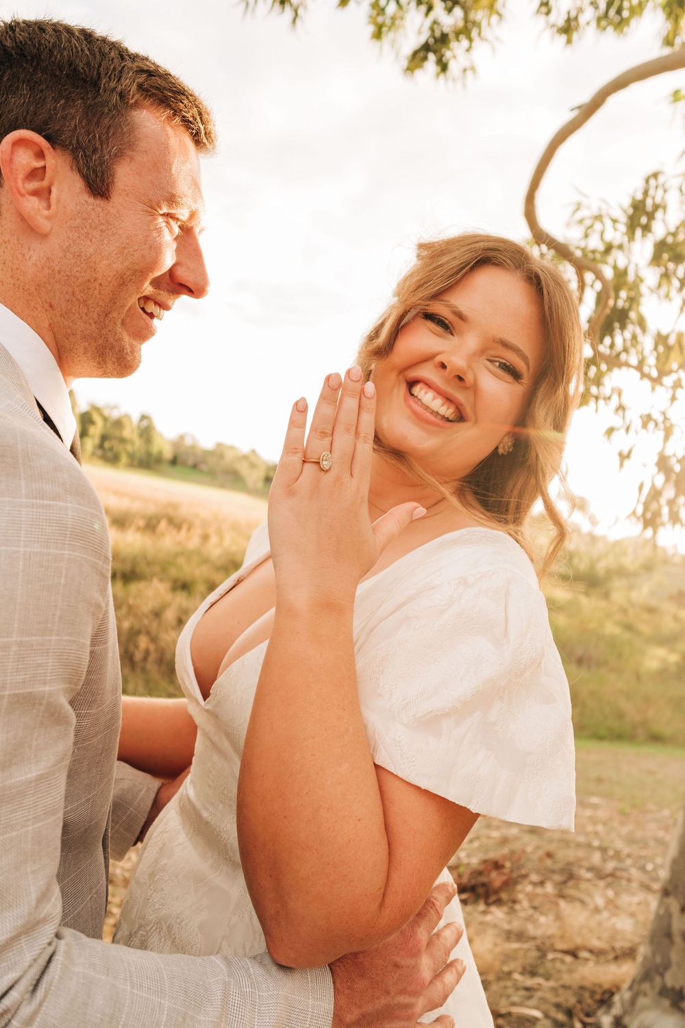Smiling bride shows off engagement ring while groom embraces her outdoors in golden sunlight near a tree.