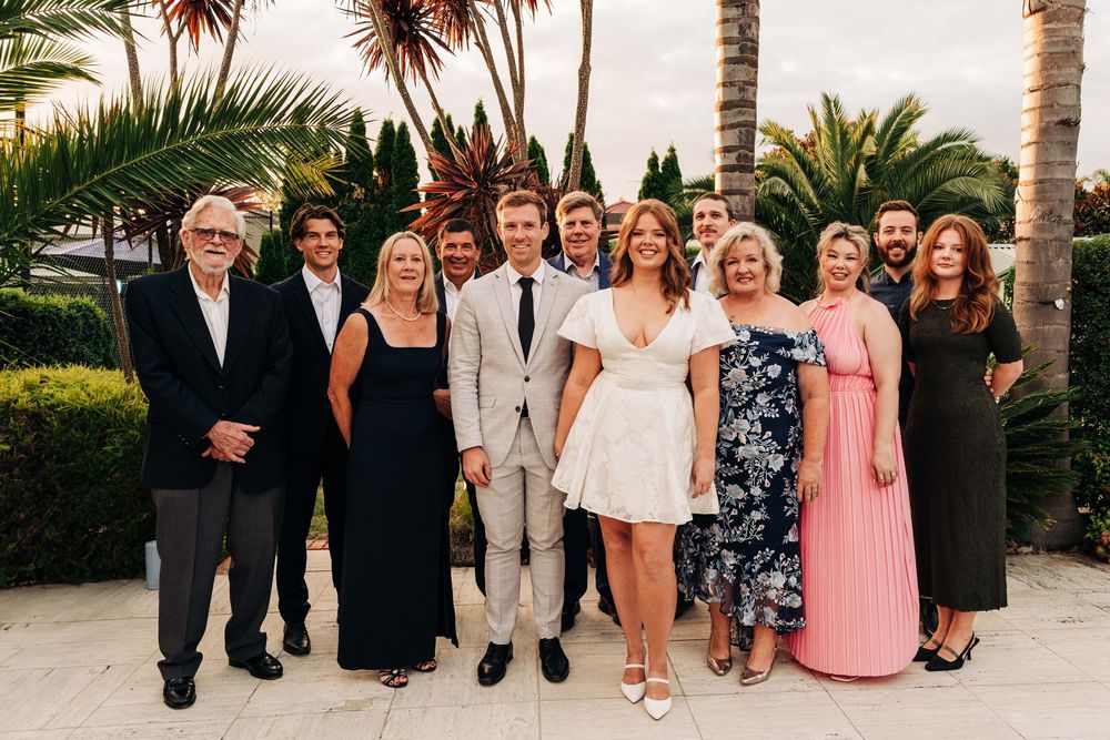 Group of elegantly dressed people posing together outdoors at a wedding celebration near palm trees.