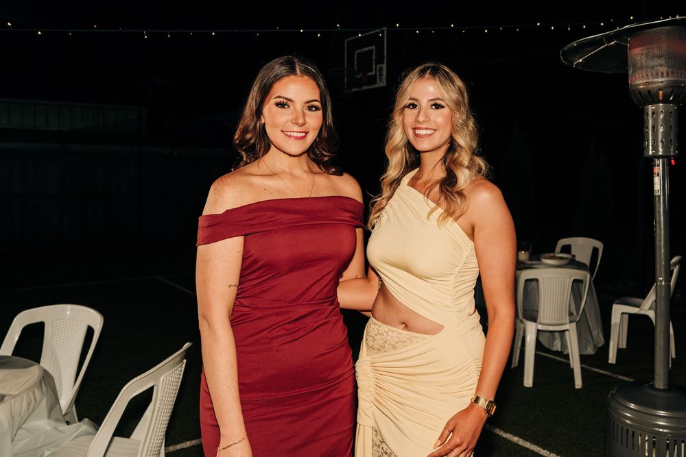 Two elegant women posing together at a formal event, wearing a burgundy off-shoulder dress and cream one-shoulder gown.