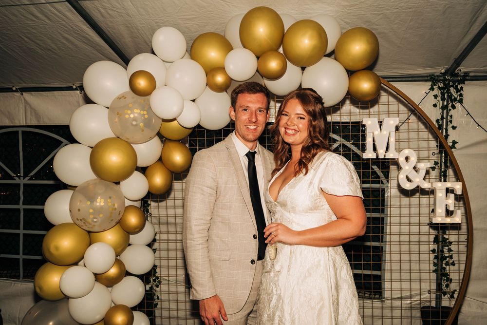 Bride and groom smiling in front of gold and white balloon arch with M&E monogram sign at wedding reception.