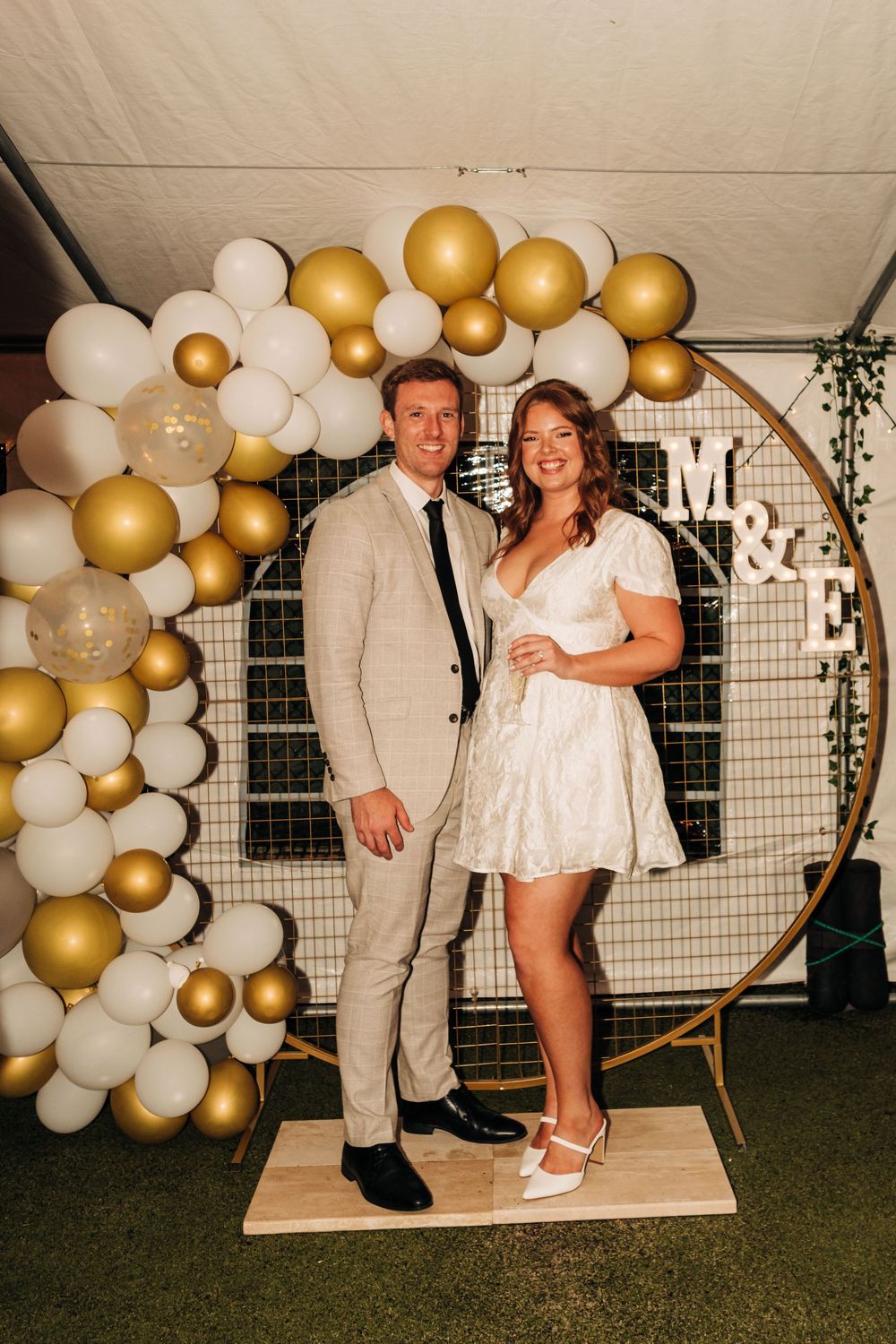 Couple posing before a gold and white balloon arch with M&E marquee letters at a wedding celebration.