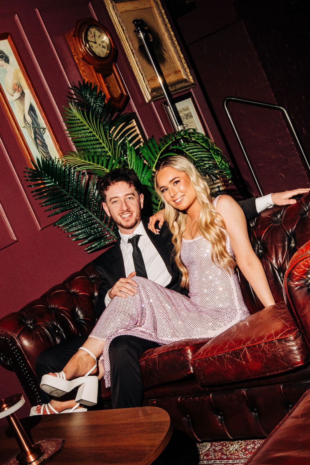 Couple in formal attire sitting on a leather sofa in an elegant dark red lounge with palm plants and vintage decor.