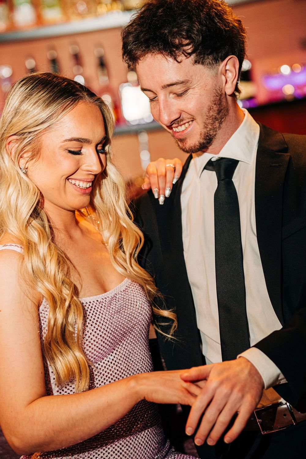 Smiling couple in formal attire sharing a joyful moment at a bar, woman in sparkly dress, man in black suit and tie.