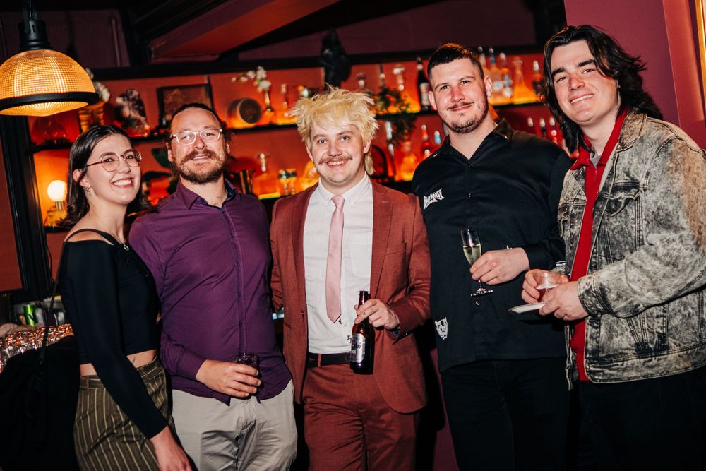 Five friends posing together at a bar, smiling for a group photo at a social event.