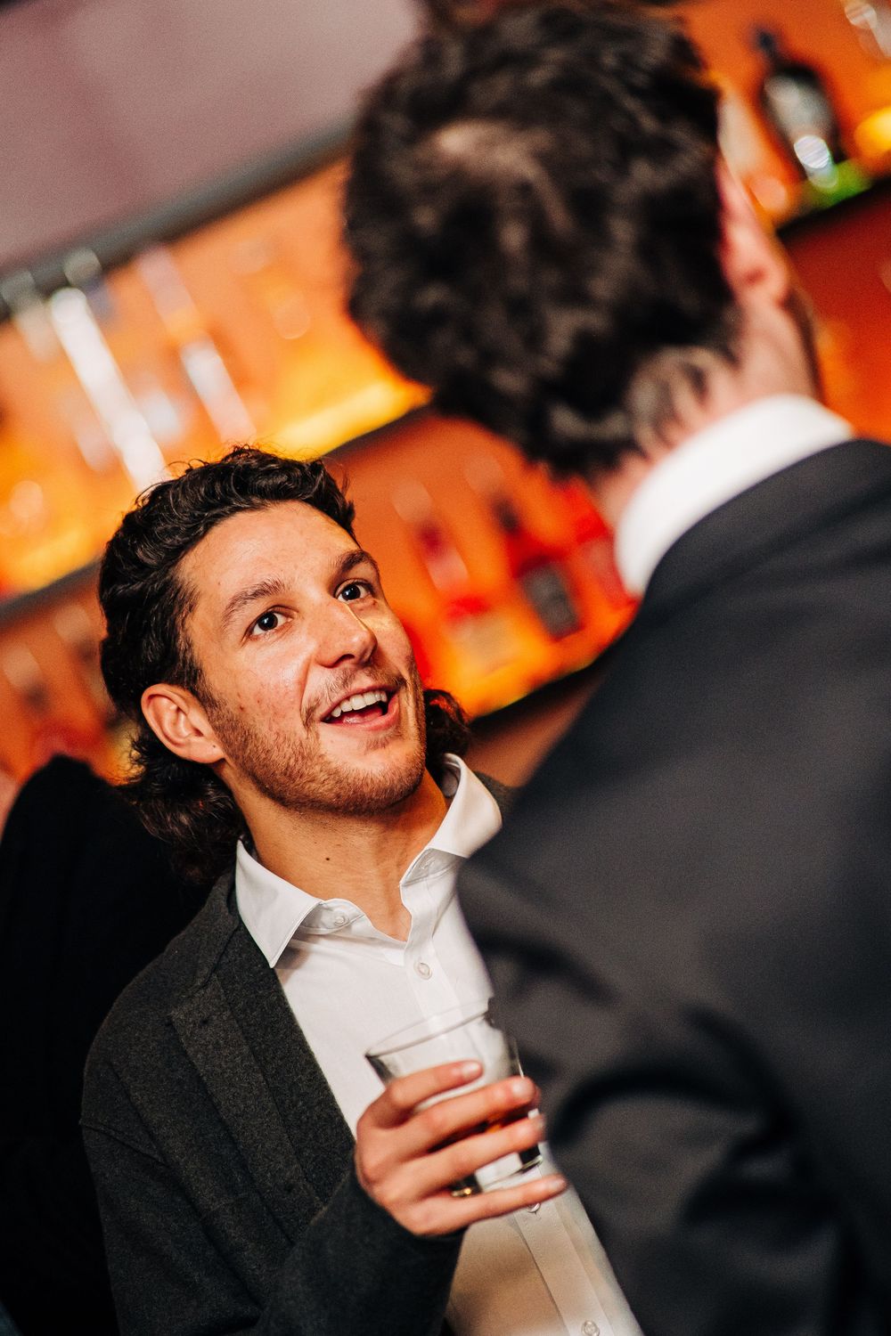 Two men in suits conversing at a bar, one smiling and holding a drink, with bottles on shelves in background.