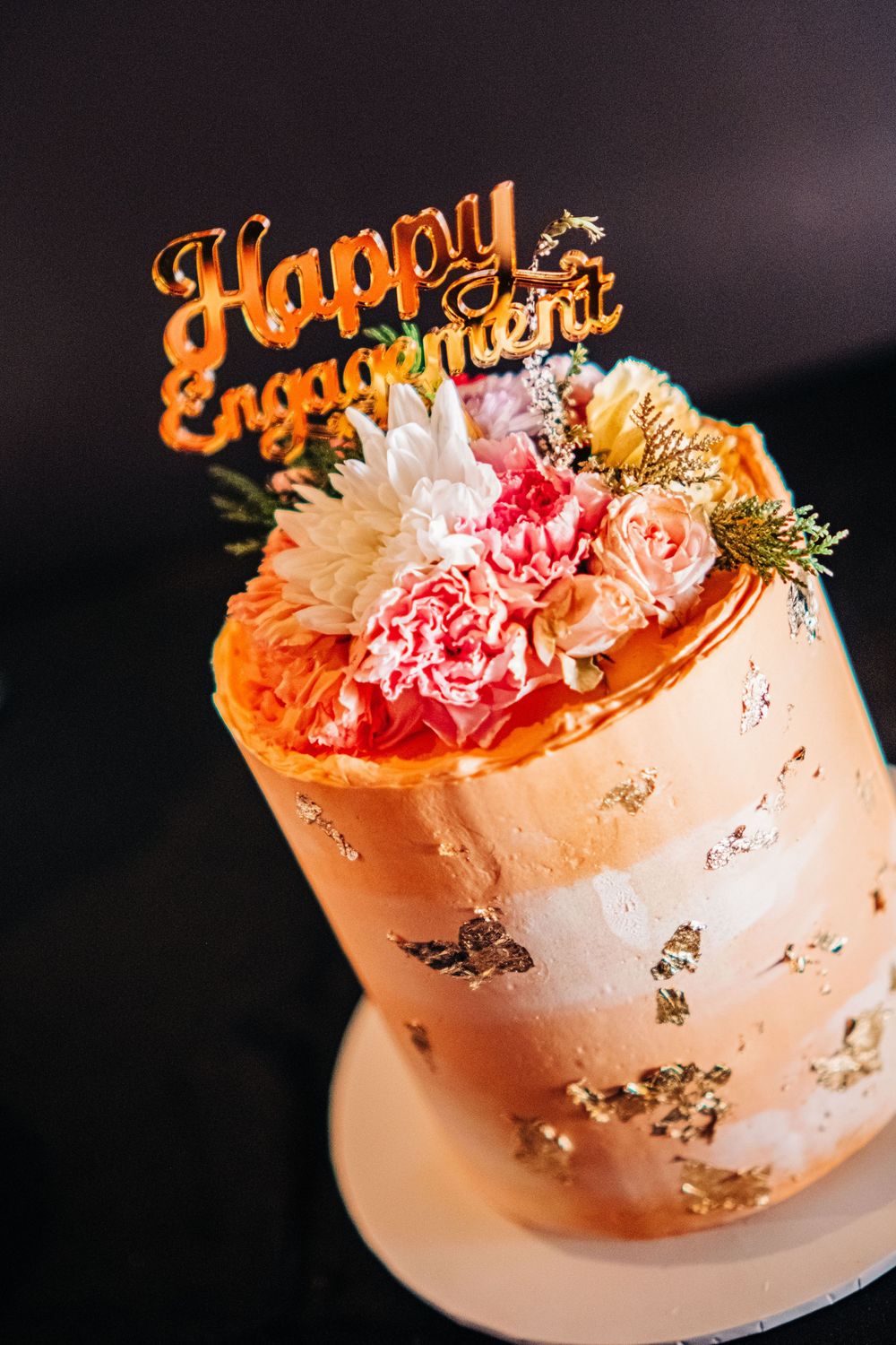 Elegant engagement cake with gold leaf, fresh pink flowers, and a gold Happy Engagement topper on dark background.