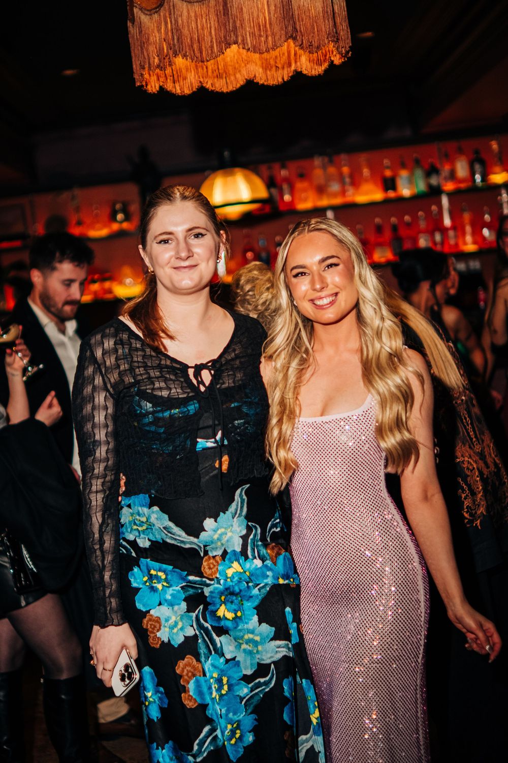Two women posing together at a bar event, one in a floral dress and one in a pink sequin gown.