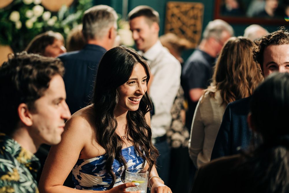 A smiling woman in a floral dress socializes at a lively indoor party gathering with guests in the background.