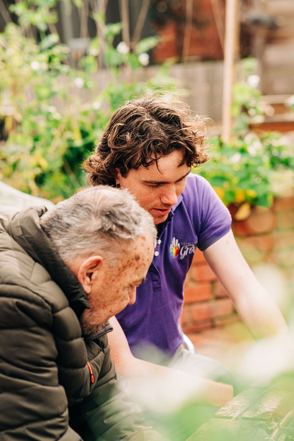Two men examining plants in a garden, one wearing a purple polo shirt outdoors.