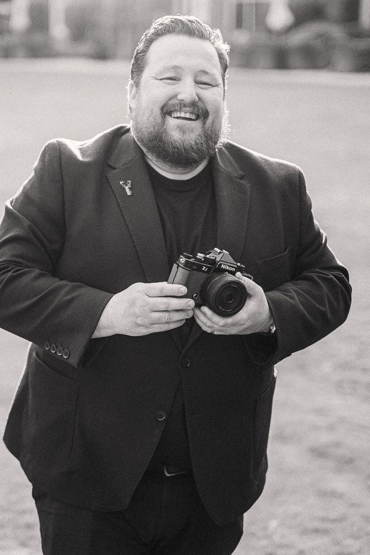 Smiling bearded man in black suit holding a camera outdoors in a black and white portrait photo.