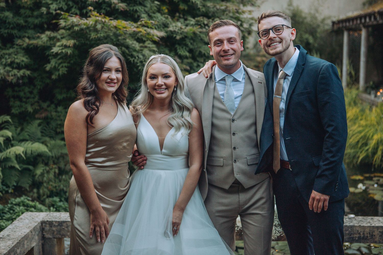 Wedding party of four smiling outdoors, bride in white gown, groom in grey suit, guests in formal attire.