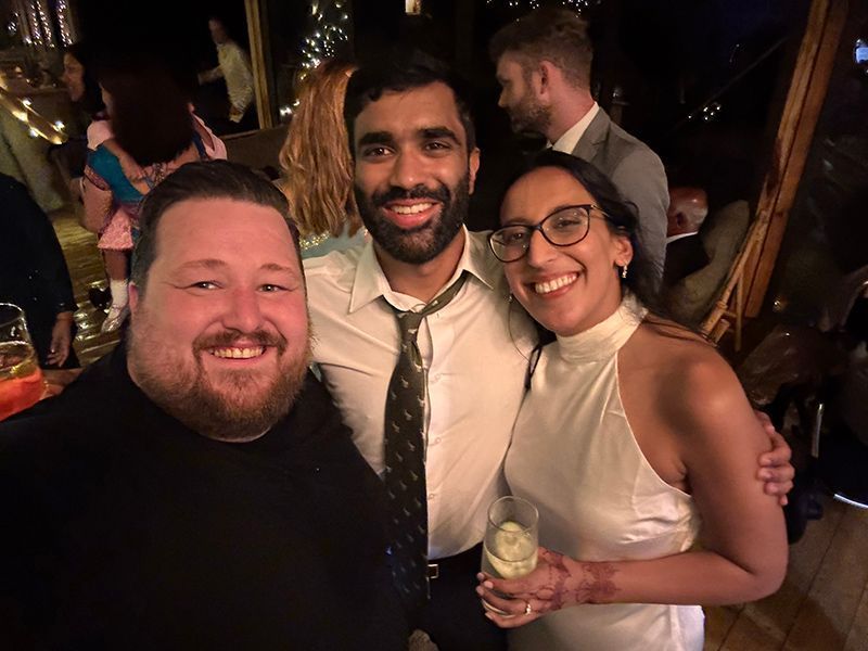 Three smiling friends pose together at an indoor evening social event, with warm ambient lighting in the background.