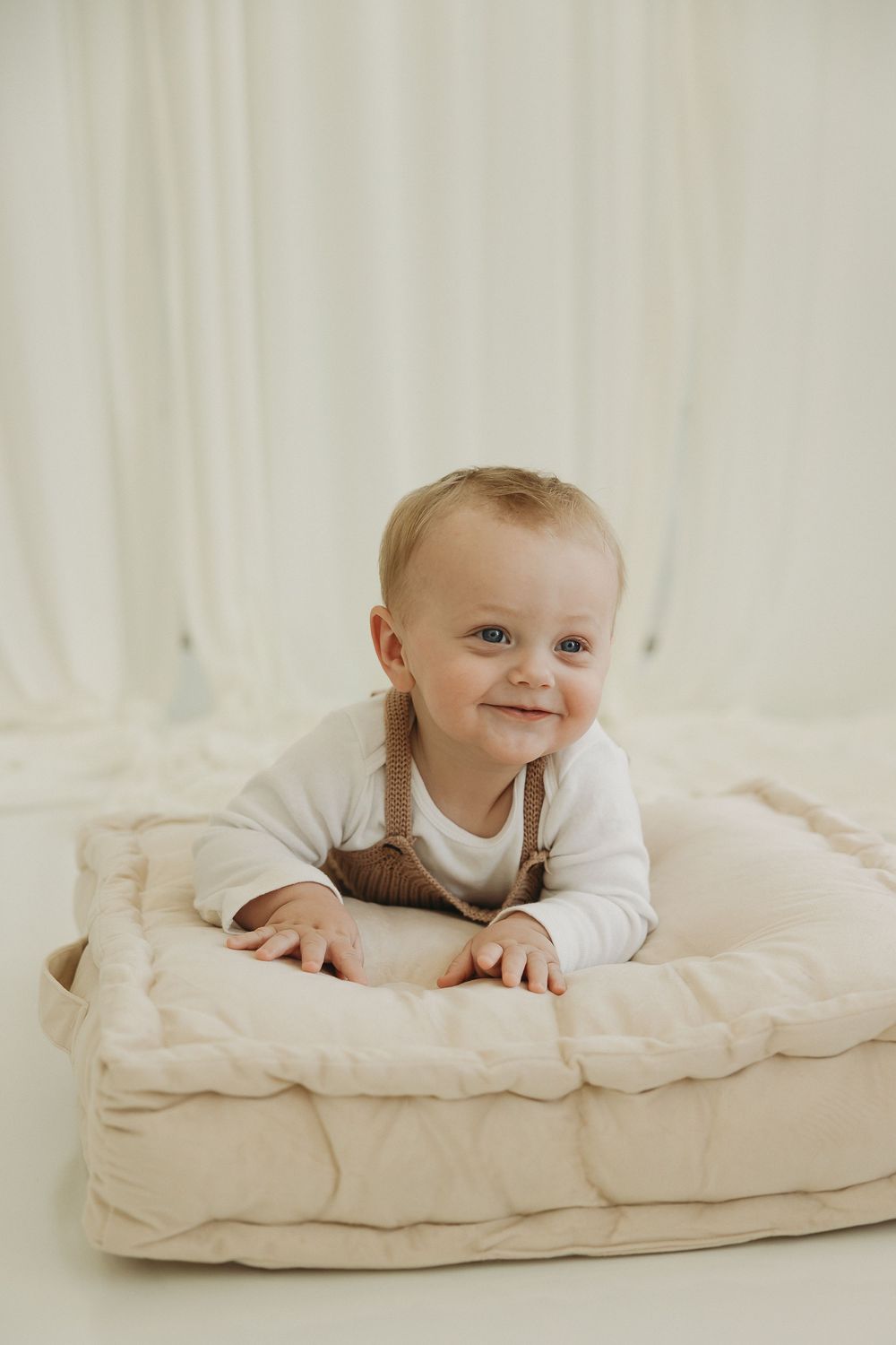 Smiling baby in white onesie and brown overalls lying on a beige quilted mat against a white background.