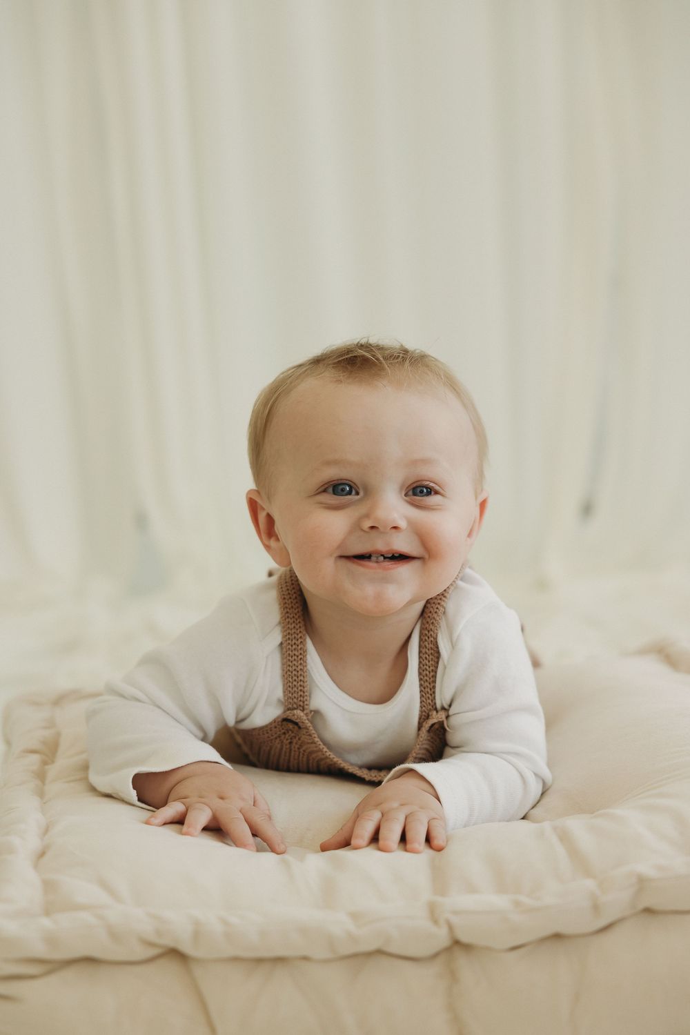 Smiling baby in white onesie and brown suspenders lying on a cream quilted blanket against a white background.