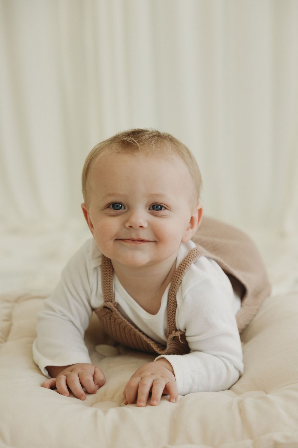 Smiling baby in brown overalls and white onesie lying on a cream blanket, looking at the camera.